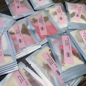 Puede incluir: Vista cenital de varios sobres pequeños y transparentes llenos de polvos de colores. Cada sobre tiene una etiqueta rosa con el texto "Mega Tea" y nombres de sabores como "Mermaid" y "Superman". Los sobres están esparcidos sobre una superficie de madera oscura.