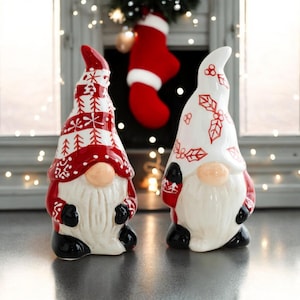 May include: Two ceramic Christmas gnomes, one with a red and white patterned hat and the other with a white hat with red holly leaves. Both gnomes have white beards and black shoes.