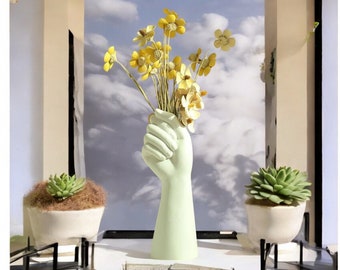 May include: A light green ceramic vase shaped like a hand holding a bouquet of yellow flowers. The vase is on a white surface with a small green succulent plant in a white pot on either side.