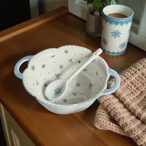 May include: A white ceramic bowl with blue handles and a matching spoon, decorated with blue snowflakes and bows. A white mug with blue snowflake designs and a knitted beige blanket are also visible.