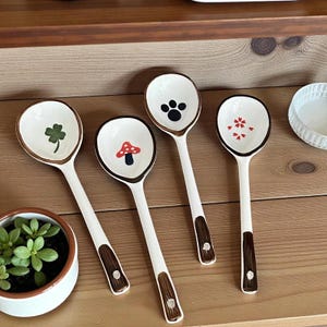 May include: Four white ceramic spoons with brown rims and handles. Each spoon bowl features a different design: a green shamrock, a red mushroom, a black paw print, and red floral accents. The spoons are arranged on a wooden surface.