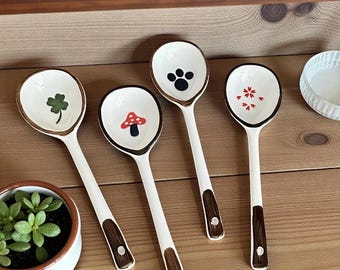 May include: Four white ceramic spoons with brown rims and handles. Each spoon bowl features a different design: a green shamrock, a red mushroom, a black paw print, and red floral accents. The spoons are arranged on a wooden surface.