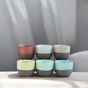 May include: Six ceramic cups with a two-tone design. The cups are arranged in two rows of three. The top row features a red, a light green, and a pink cup. The bottom row features a yellow, a light blue, and a turquoise cup. All cups have a dark gray base.