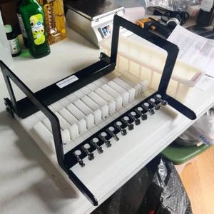 May include: A black and white soap cutter with a series of vertical white dividers. The cutter has a black frame and a row of silver-colored levers. The cutter is on a white surface.