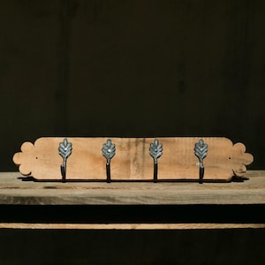 May include: A wooden wall-mounted coat rack with four hooks. The hooks are made of black metal and have a leaf design. The rack is made of natural wood with a light brown finish.