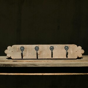 May include: A rustic wooden coat rack with four metal hooks. The light-colored wood has a natural grain and is shaped with decorative floral edges. The hooks are dark metal and have a flower design.