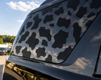 Op de afbeelding: Een achterste zijruit van een zwart voertuig met een matzwarte koeienvlekkensticker. Het patroon bestaat uit diverse onregelmatige, afgeronde vormen die op het glasoppervlak zijn aangebracht en contrasteren met het glanzend zwarte frame.