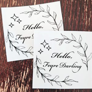 May include: Two white cards with black ink. Each card features a wreath of leaves and the text "Hello, Feyre" with stars. The second card has the text "Darling" after "Feyre".