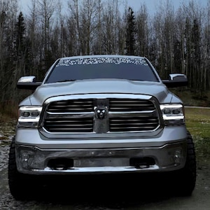K&ouml;nnte beinhalten: Ein silberner Pickup-Truck mit einem markanten Chromk&uuml;hlergrill und zentralem Widder-Emblem, rechteckigen Scheinwerfern mit integrierten LED-Streifen und einem Windschutzscheibenaufkleber mit wei&szlig;em Leopardenmuster am oberen Rand.