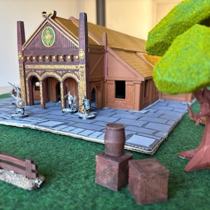 May include: A miniature fantasy diorama featuring a brown wooden building with gold accents, a green and brown tree, and several miniature figures. The scene is set on a green surface with a gray stone-like base. Additional elements include a barrel and crates.
