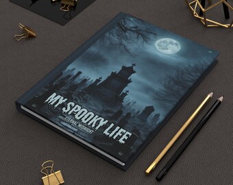 May include: A hardcover journal with a spooky graveyard scene under a full moon. The cover reads "MY SPOOKY LIFE" with "Eternal Midnight" below. Gold and black pencils and gold binder clips are scattered around the journal.