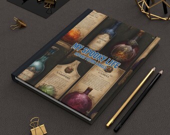 May include: A hardcover journal with the text "My Spooky Life, Arcane Potion Labels" on the cover. The cover features illustrations of potion bottles with labels. Gold and black pencils and binder clips are on the dark gray surface.