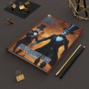 Peut inclure: Un journal à couverture rigide avec une bordure marron et le texte "MY SPOOKY LIFE DAPPER DEAD". La couverture présente une illustration numérique de deux squelettes en tenue de soirée. Des crayons dorés et noirs et des trombones dorés sont également visibles.
