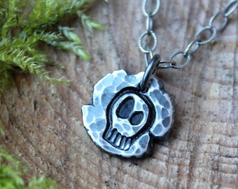 May include: A silver necklace with a hammered metal skull pendant. The pendant has a textured surface and a simple skull design.