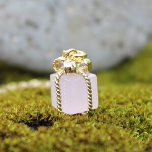 May include: A gold-colored pendant shaped like a gift box, with a pink, rectangular body and a gold bow on top. The pendant hangs from a twisted gold chain. The background is blurred, with green moss and a light-colored rock.