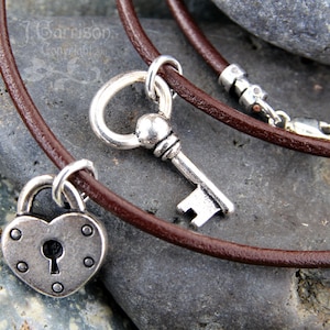 以下が含まれることがあります： 銀色のハート型の南京錠と銀色の鍵のチャームが付いた、茶色の革紐のネックレス。ネックレスは灰色の石の背景の上に置かれています。