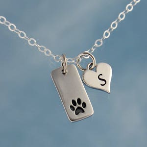 May include: Silver necklace with a small rectangular tag featuring a paw print and a heart charm with the letter S.