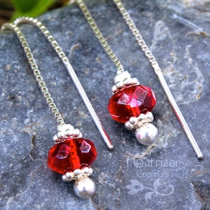 May include: A pair of dangle earrings featuring faceted red glass beads and silver-tone metal accents. The earrings have a long, thin chain that threads through the ear. The design includes small silver beads and a larger, round bead at the bottom.