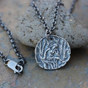 Op de afbeelding: Een zilveren ketting met een ronde hanger met een gedetailleerd reliëf van twee figuren in een boslandschap.