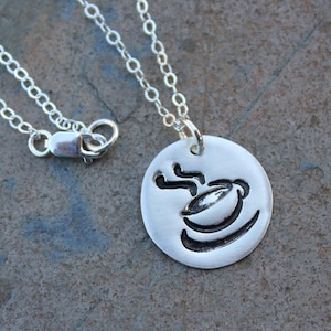Könnte beinhalten: Eine silberne Halskette mit einem runden Anhänger, der ein stilisiertes Bild einer Kaffeetasse mit aufsteigendem Dampf zeigt.