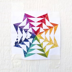May include: A colorful, geometric quilt block featuring a starburst design. The star is composed of various triangles in a rainbow of colors, including red, orange, yellow, green, blue, and purple. The block is set against a white background.
