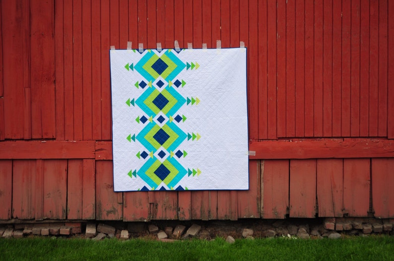 May include: A white quilt with a geometric pattern in shades of blue, green, and yellow. The quilt is hanging on a red barn.