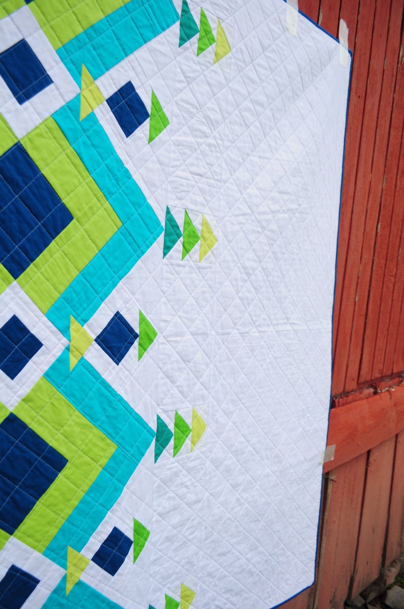 May include: A white quilt with a geometric pattern in shades of blue, green, and yellow. The quilt is hanging on a red wooden wall.