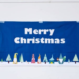 Peut inclure: Une banni&egrave;re bleue avec le texte "Merry Christmas" en lettres blanches. La banni&egrave;re est d&eacute;cor&eacute;e d'une rang&eacute;e de gnomes et d'arbres color&eacute;s.