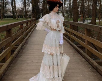 Puede incluir: Una mujer con un vestido blanco con ribetes de encaje y un sombrero marrón se encuentra de pie en un puente de madera. El vestido tiene una falda de capas y un corpiño ajustado. La mujer sostiene una sombrilla blanca.