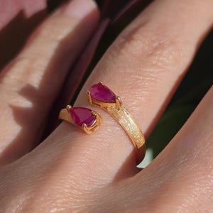 Puede incluir: Un anillo abierto de oro con un acabado mate y texturizado en la banda. El anillo está adornado con dos piedras preciosas de color rosa intenso en forma de pera, cada una sujeta en un engaste de garras en los extremos abiertos.