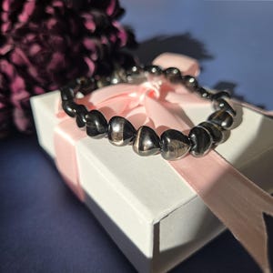 Puede incluir: Una pulsera oscura con cuentas, con una mezcla de cuentas redondeadas y oblongas, descansa sobre una caja de regalo blanca atada con una cinta rosa. Las cuentas de la pulsera tienen un brillo metálico. Un arreglo floral morado oscuro está en el fondo.