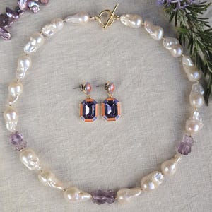 Op de afbeelding: Een ketting met onregelmatige witte barokparels afgewisseld met kleine paarse gefacetteerde kralen en een goudkleurige knevelsluiting, vergezeld van een paar bijpassende oorbellen met rechthoekige paarse stenen in oranje en goudkleurige randen.