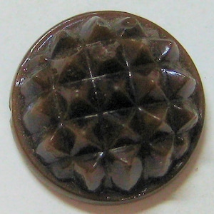 May include: A dark brown, round button with a raised, geometric design. The button has a textured surface with a pattern of small, pointed shapes.