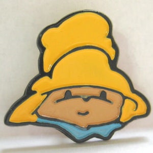 May include: A yellow enamel pin with a cartoon character wearing a yellow hat and a blue scarf. The character has a brown face and black eyes.