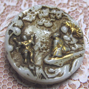 May include: A round, off-white button with a relief design featuring two figures and foliage in a landscape. The figures and some details are accented with gold. The button has a slightly aged appearance.