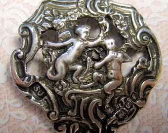 May include: Ornate silver-toned brooch featuring a detailed relief of two cherubic figures amidst scrolling foliage. The brooch has a decorative, openwork border with floral accents and is likely vintage.