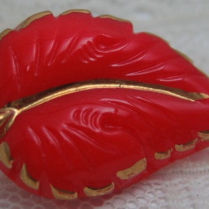 May include: A red plastic button shaped like a leaf with gold trim. The leaf has a raised design.