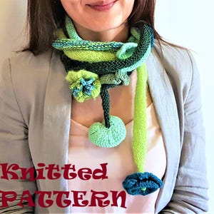 Op de afbeelding: Een handgebreide sjaal in groen, blauw en zwart, met bloemen- en pompomdetails. De sjaal heeft een uniek ontwerp met een lang, vloeiend element. De tekst "Knitted Pattern" is zichtbaar. Gedragen door een persoon in een lichtgrijze blazer.