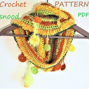 May include: A handmade crochet snood in shades of yellow, orange, and green, adorned with dangling pom-poms and circular accents. The text "Crochet snood PATTERN PDF" is visible. The snood is displayed on a wooden hanger.