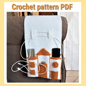 Könnte beinhalten: Ein weiß-oranger Häkel-Organizer mit Taschen für Fernbedienungen und ein Telefon. Der Organizer ist so konzipiert, dass er über der Armlehne eines Sofas hängt. Der Text "Crochet pattern PDF" befindet sich oben im Bild.