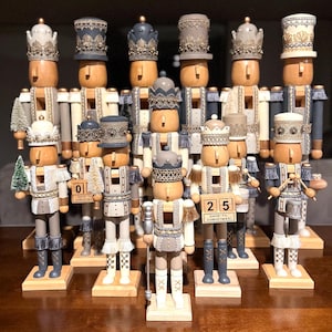 May include: A collection of 12 wooden nutcracker figurines, each with a unique design and painted in shades of gray, white, and gold. The nutcrackers are standing on wooden bases and feature a variety of details, including hats, sashes, and instruments. Some of the nutcrackers have a small sign with the number of days until Christmas.