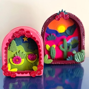 May include: Two decorative shadow box art pieces. One is pink with a floral and landscape design, the other is maroon with a desert landscape. Both have a curved arch shape and are decorated with roses.