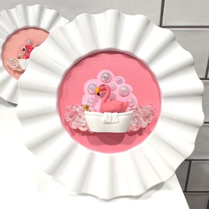 May include: Two decorative wall art pieces. Each features a pink background with a flamingo in a bathtub. The flamingo is surrounded by bubbles and clear embellishments. The art is framed with a white, ruffled edge.