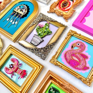 May include: A collection of small, ornate frames containing beaded art pieces. The subjects include a jellyfish, a cactus, a flamingo, a lobster, a monstera leaf, a peach, and a pink bow. Each piece features intricate beadwork in vibrant colors.