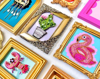 May include: A collection of small, ornate frames containing beaded art pieces. The subjects include a jellyfish, a cactus, a flamingo, a lobster, a monstera leaf, a peach, and a pink bow. Each piece features intricate beadwork in vibrant colors.