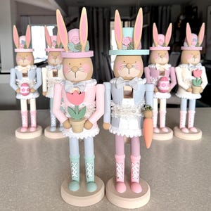 May include: A set of six pastel-colored wooden bunny nutcrackers. Each bunny is wearing a different hat and holding a different Easter-themed accessory. The bunnies are standing on a light brown surface.