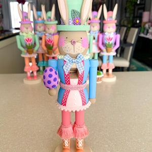 May include: A pastel blue and pink Easter bunny nutcracker with a green hat, holding a purple and white polka dot egg. The nutcracker is standing on a cream-colored base.