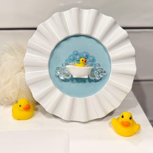 May include: A white, ruffled-edge frame with a light blue center features a yellow rubber duck in a bathtub with bubbles. Two additional yellow rubber ducks are placed on the white surface, creating a playful, bath-themed decorative item.