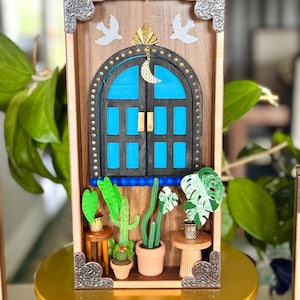 May include: A wooden shadow box featuring a blue arched window with gold accents, a crescent moon charm, and two white dove cutouts. Miniature potted plants, including cacti and a monstera, are arranged below the window. The frame has decorative silver corner pieces.