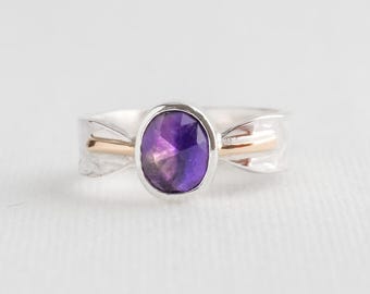 May include: A silver ring with a gold band and a large, oval-cut purple gemstone. The ring's design features a delicate, flowing pattern on the sides. The gemstone is the focal point of this elegant piece of jewellery.